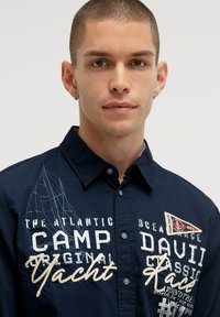 Marineblaue Baumwollhemd mit Knopfverschluss, bestickt mit Grafiken, einem Segelboot-Design und kontrastierendem Text auf der Vorderseite.