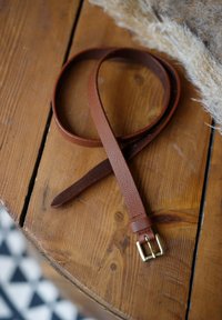 Brown leather belt on a wooden table, featuring a gold metal buckle. The belt has a textured finish and tapering ends.
