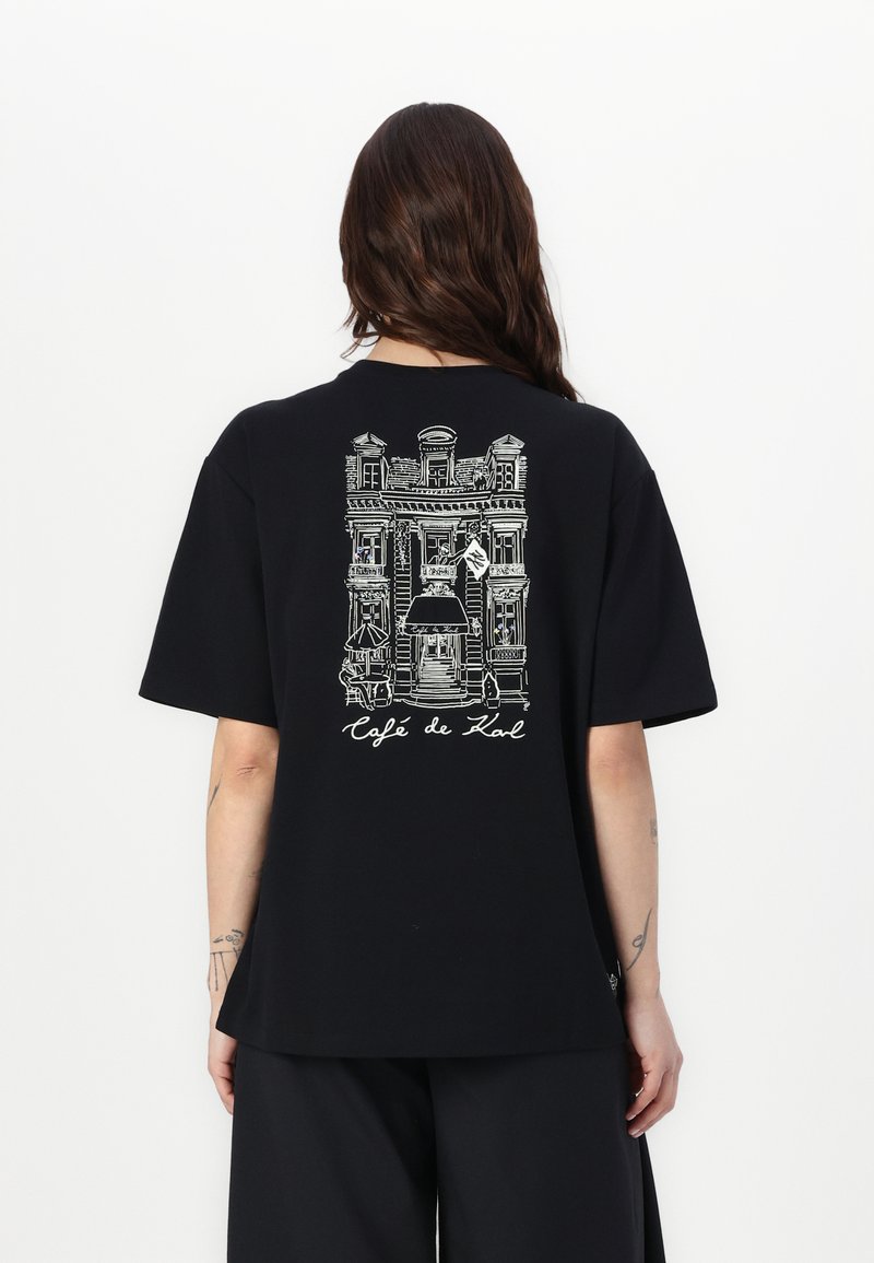 Schwarzes, übergroßes T-Shirt mit einer weißen Linienzeichnung der Fassade eines Cafés und dem Text "Café de Lowl" auf der Mitte des Rückens, kurze Ärmel.