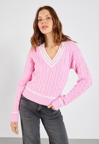 Roze kabelgebreide trui met een v-hals en een contrasterende witte streep bij de kraag en de zoom. De mouwen hebben een ribpatroon.