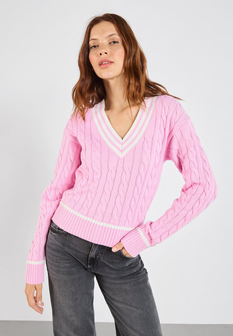 Roze kabelgebreide trui met een v-hals en een contrasterende witte streep bij de kraag en de zoom. De mouwen hebben een ribpatroon.