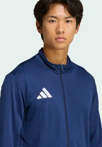 Ναυτικό μπουφάν με φερμουάρ κατασκευασμένο από μαλακό ύφασμα, με ψηλό γιακά και λευκό λογότυπο Adidas στην αριστερή πλευρά.