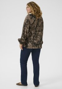 Femeie cu păr ondulat blond, îmbrăcată cu o bluză lungă cu model maro, blugi negri și balerini maro, stând de profil pe un fundal simplu.