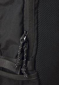 Črn nahrbtnik iz materiala s teksturirano tkanino in mrežnimi detajli. Ima dvojne zadrge in trdne vrvi z odsevnimi detajli.