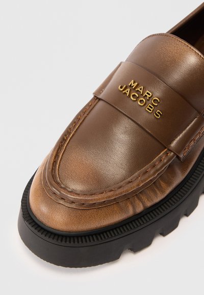 Hnědé kožené mokasíny s kulatou špičkou, s zlatým logem "Marc Jacobs" a silnou černou gumovou podrážkou s texturovaným povrchem.