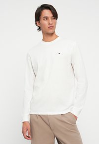 Mand med mellemlangt mørkt hår iført en hvid langærmet skjorte og beige bukser, der kigger let til siden mod en ensfarvet baggrund.