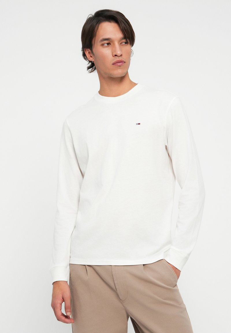 Hombre con cabello oscuro de longitud media, vistiendo una camisa blanca de manga larga y pantalones beiges, mirando ligeramente de lado contra un fondo sencillo.