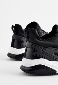 Czarno-białe sportowe sneakersy widziane z tyłu, z fakturowanymi podeszwami, uchwytami do zakupu oraz smukłymi syntetycznymi nakładkami.