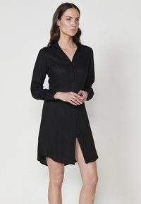 Rochie neagră cu nasturi, confecționată dintr-un material ușor, având guler, mâneci lungi și o fanta adâncă. Finish texturat și croială lejeră.