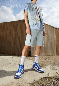 Chaleco de mezclilla azul claro con bolsillos, combinado con una camiseta gráfica azul claro, pantalones cortos de mezclilla a juego y zapatillas altas en azul y blanco.