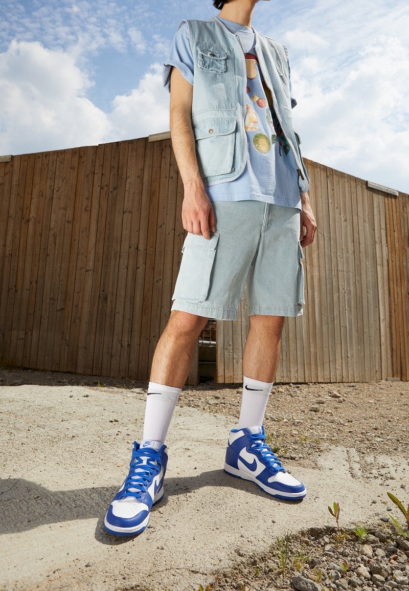 Chaleco de mezclilla azul claro con bolsillos, combinado con una camiseta gráfica azul claro, pantalones cortos de mezclilla a juego y zapatillas altas en azul y blanco.