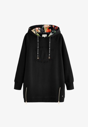 Vrouw staat op blote voeten en draagt een lange zwarte hoodie met een patroon op de capuchon, met haar rug naar een effen lichte achtergrond gekeerd.