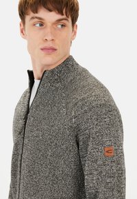 Grauer Strickpullover mit hohem Kragen, Reißverschlussverschluss, strukturiertem Muster und braunem Lederlogo-Patch am linken Arm.