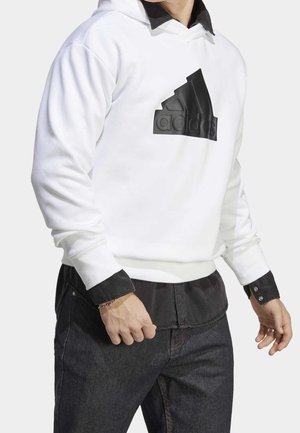 Weißer Hoodie mit einem schwarzen, gummierten Logo, ausgestattet mit einer Kängurutasche und gerippten Bündchen. Kombiniert mit dunklen Jeans und einem schwarzen Shirt darunter.