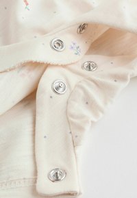 Tissu beige clair avec de petits motifs floraux colorés et des fermetures à pression métalliques sur le bord, présentant une texture douce et une légère élasticité.