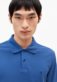 Blaues Poloshirt aus strukturiertem Stoff, mit einem Kragen, drei Knöpfen und einem kleinen gestickten Logo auf der linken Brust.