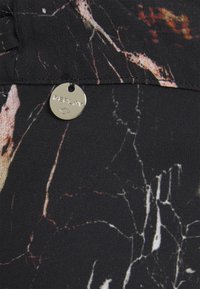 Tissu noir avec un léger motif marbré et une petite étiquette métallique ronde argentée gravée du mot « REPLAY », attachée près d'une couture.