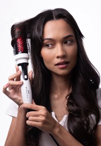 Eine weiße beheizte Stylingbürste mit einem roten Netzfass, die von einer Person gehalten wird, verfügt über ein digitales Display und schwarze Akzente. Haare werden gestylt.