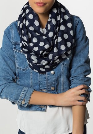 Femme portant un foulard bleu marine à pois blancs sur une veste en jean bleue et une chemise blanche, les bras croisés à la taille.