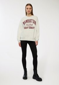 Felpa grigio chiaro con la scritta "MANHATTAN" e "EAST COAST" stampate in rosso scuro. Abbinata a leggins neri e stivali neri con lacci.