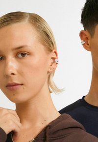 Nærbillede af to personer iført sølv ørekæder, den ene med kort blont hår og en brun hættetrøje, den anden med kort mørkt hår og en sort skjorte.