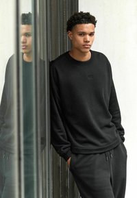 Sort sweatshirt med ribbet halsudskæring, der har subtil branding. Parret med matchende sorte sweatpants, glat tekstur, afslappet pasform.