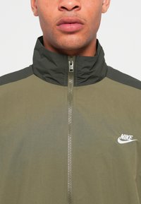 Moški v olivno zeleni in temno zeleni Nike jopici z zadrgo in visokim ovratnikom ter belim logotipom na prsih.