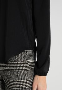 Schwarzes Langarmshirt mit leicht abgerundetem Saum, kombiniert mit karierten schwarz-weißen Hosen aus einem strukturierten Stoff.