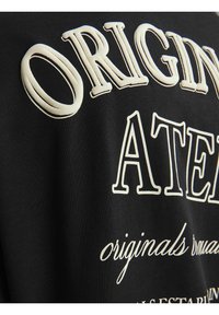 Sort t-shirt med hævet, cremet tekst, herunder "ORIGINAL" og "ATELIER", lavet af glat bomuldsstof.