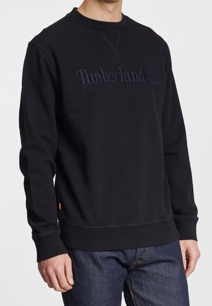 Felpa nera Timberland con colletto, polsini e orlo a coste indossata da una persona in jeans blu scuro, con logo sul petto e testo.