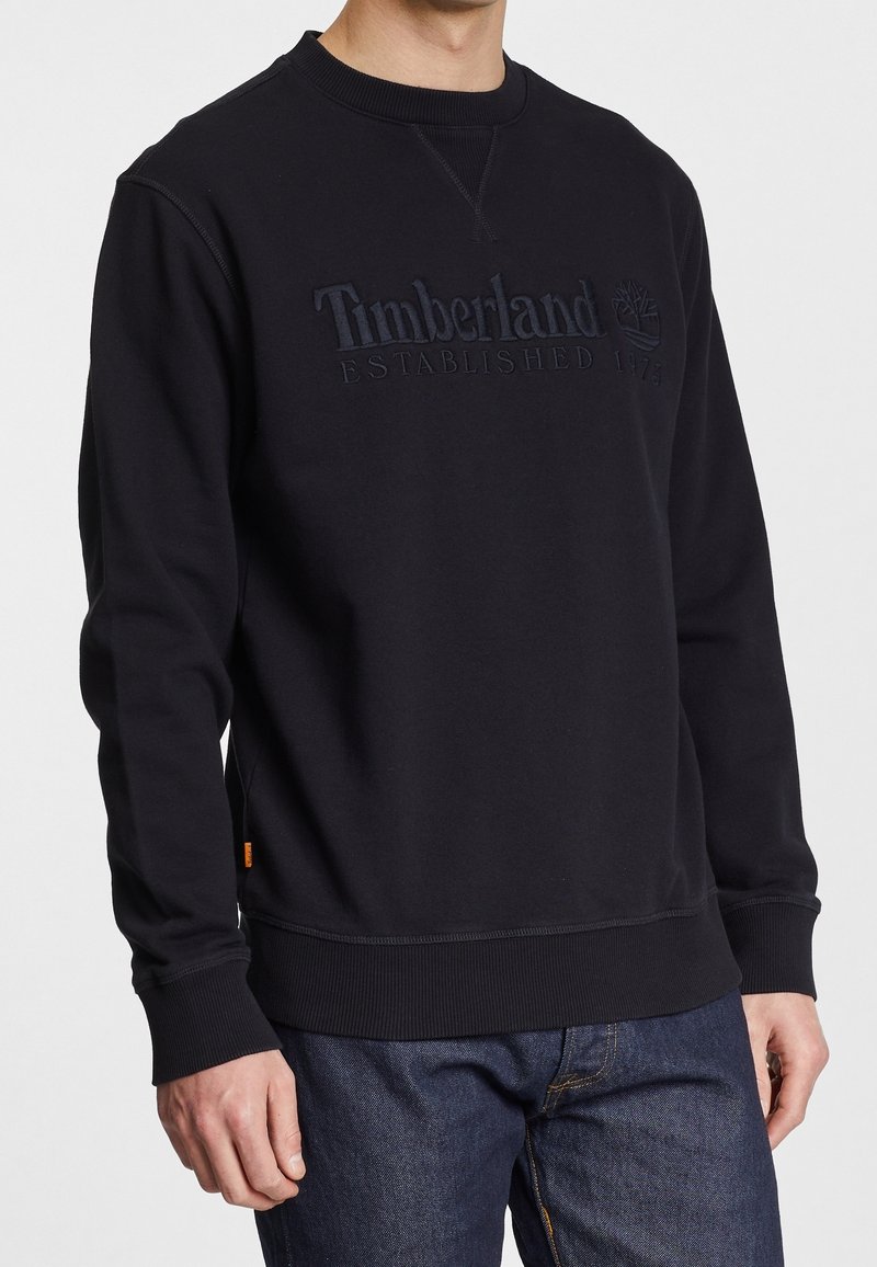 Svart Timberland-tröja med ribbad krage, manschetter och nederkant, bärs av person i mörkblå jeans, med bröstlogotyp och text synlig.