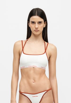 Kobieta ubrana w biały i rdzawego koloru braletę Calvin Klein oraz pasujące do niej majtki bikini stoi na jednolitym białym tle.