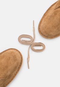 Zapatos deslizantes de ante beige con una textura suave. Dos cordones beige están enrollados en el centro. Forma redonda simple y sin herrajes visibles.