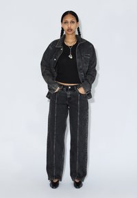 Veste en denim noir et jeans assortis à coupe décontractée, associés à un crop top noir et des chaussures noires. Des bijoux argentés viennent parfaire la tenue.