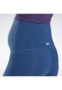 Leggings azuis de cintura alta com uma textura suave e um brilho sutil, apresentando uma blusa roxa, costuras planas e um pequeno logótipo na cinta.