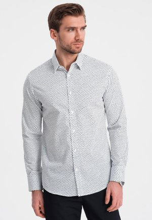Camicia bianca con piccoli pois neri, caratterizzata da un colletto classico e maniche lunghe. Il modello la indossa con pantaloni scuri.