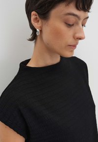 Schwarzes, geripptes Oberteil mit hohem Kragen und kurzen Ärmeln. Das Model trägt glänzende silberne geometrische Ohrringe. Glatt gewebter Stoff.