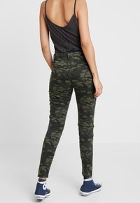 Zelené kalhoty s kamuflážním vzorem vyrobené z bavlny, přiléhavý střih, zúžené nohavice, v kombinaci s modrými plátěnými teniskami, zobrazené zezadu.