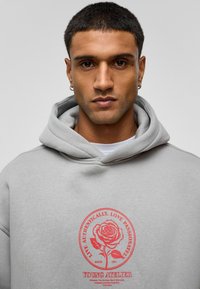 Grijze hoodie van zachte stof, met een rode rozenafbeelding en tekst op de voorkant en achterkant. Bevat een grote capuchon en geribde manchetten.