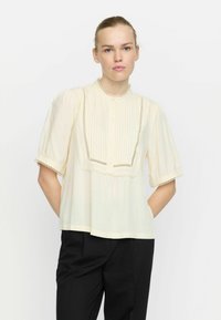 Cremfarbene Bluse mit kurzen Puffärmeln, gefältelten Details, Spitzenbesatz und rundem Halsausschnitt, kombiniert mit schwarzen Hosen.