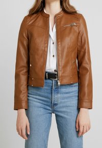 Brun læderjakke med rund halsudskæring, lynlås foran og sidelommer. Bærer den over en hvid skjorte og kombineret med blå jeans.