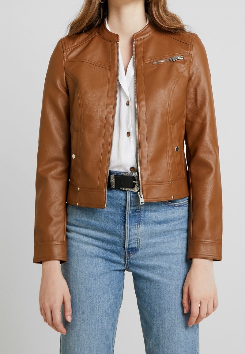 Brun læderjakke med rund halsudskæring, lynlås foran og sidelommer. Bærer den over en hvid skjorte og kombineret med blå jeans.