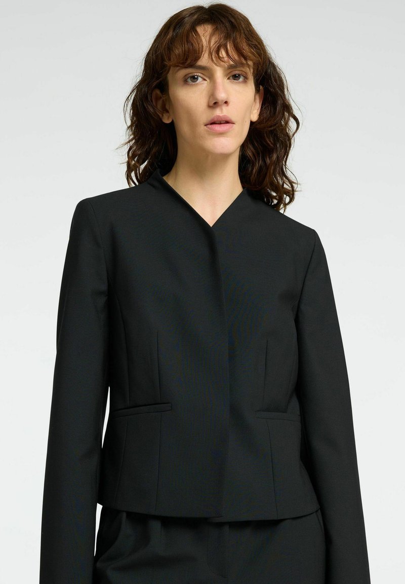 Frau mit lockigem braunem Haar, die einen taillierten schwarzen Blazer trägt und vor einem schlichten hellen Hintergrund steht, frontal ausgerichtet.