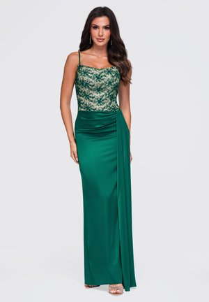 Femme portant une robe de soirée vert émeraude avec un corsage en dentelle, des bretelles fines et un détail de jupe drapée, debout devant un fond clair uni.