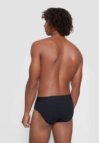 Schwarze Baumwollslips mit glatter Textur, elastischem Taillenbund und taillierter Silhouette, von hinten gegen einen hellen Hintergrund betrachtet.