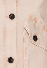 Tessuto di denim beige chiaro con bottoni neri e una tasca a pattina cucita su un capo, che mostra dettagliata texture e cuciture.