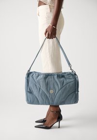 Modrá džínová taška na rameno se shromážděným vrškem, stříbrným detailem loga a nastavitelným popruhem. V kombinaci s krémovými kalhotami a černými špičatými podpatky.