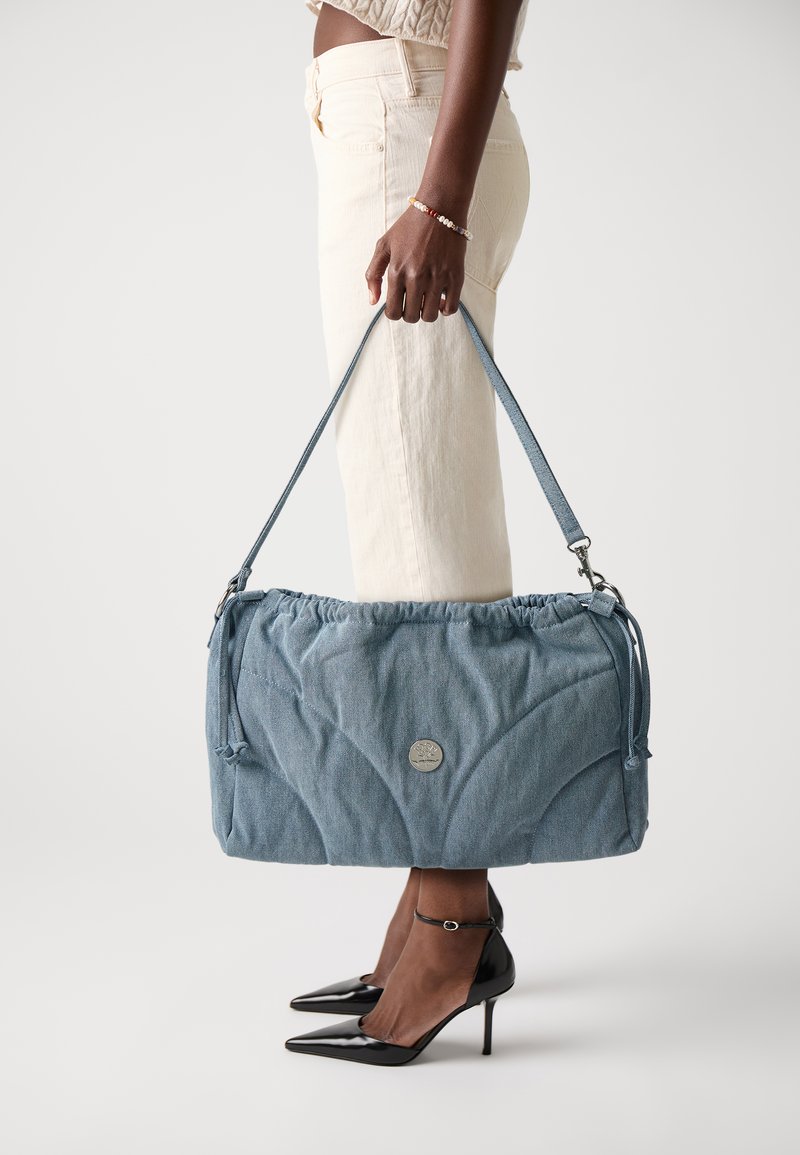 Modrá džínová taška na rameno se shromážděným vrškem, stříbrným detailem loga a nastavitelným popruhem. V kombinaci s krémovými kalhotami a černými špičatými podpatky.
