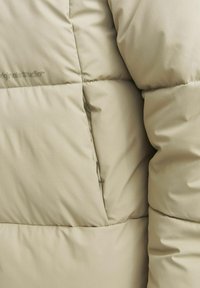 Beige puffjakke sektion med quiltet tekstur, der viser glat stof og en subtil glans. Har syning detaljer og en sidelomme.