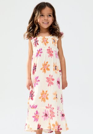 Junges Mädchen mit schulterlangem lockigem Haar, trägt ein ärmelloses weißes Kleid mit rosa, orangefarbenen und roten Blumenmustern, lächelnd und stehend.